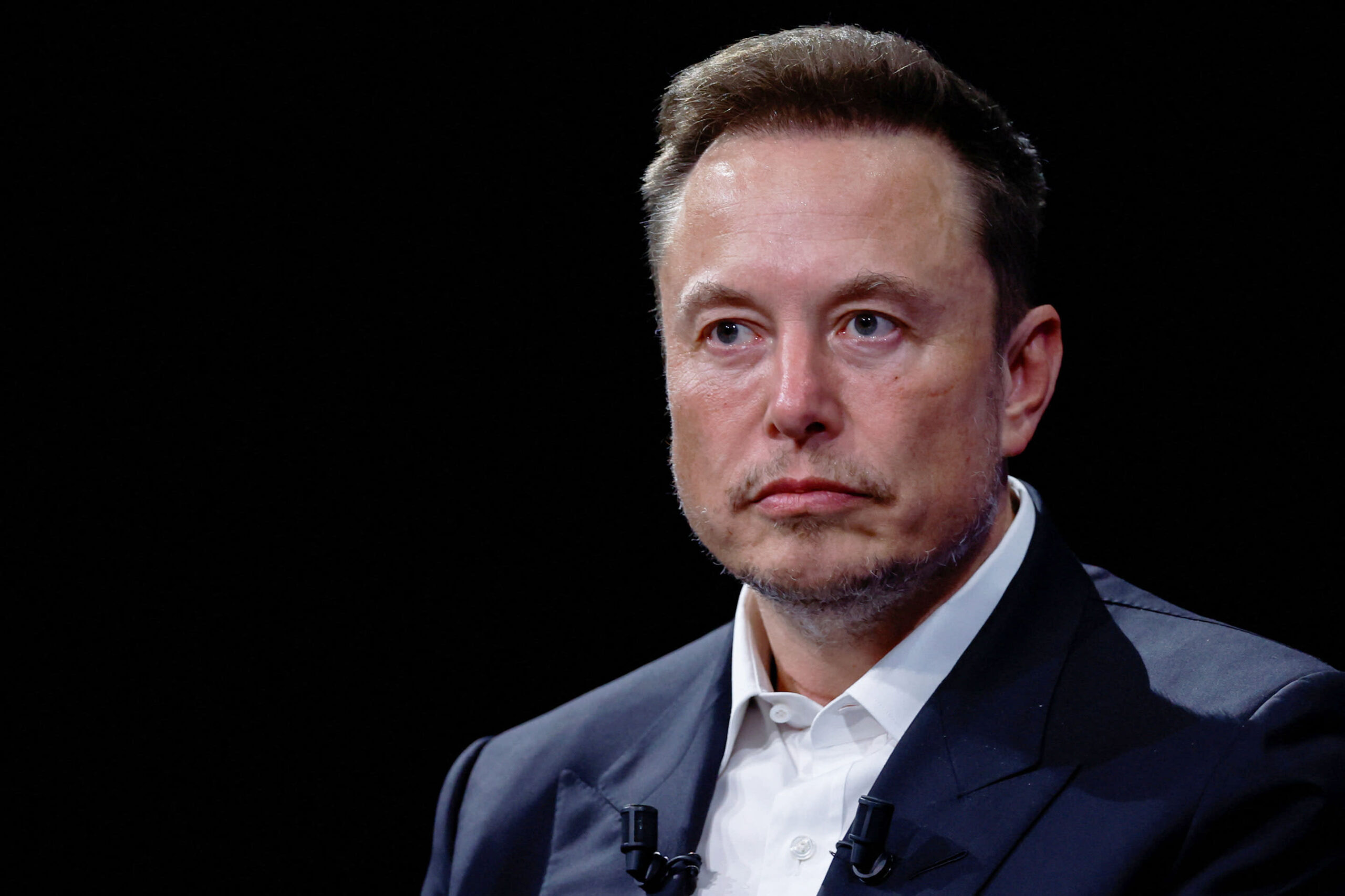 image shows a headshot of Elon Musk wearing a suit staring at the camera.