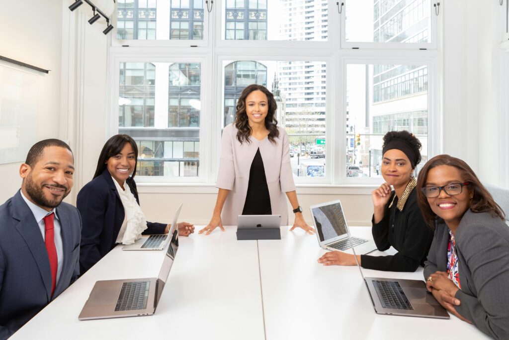 Image shows five professionally dressed people seated around a white conference table with a view of a downtown area. Each person has a laptop indent of them.