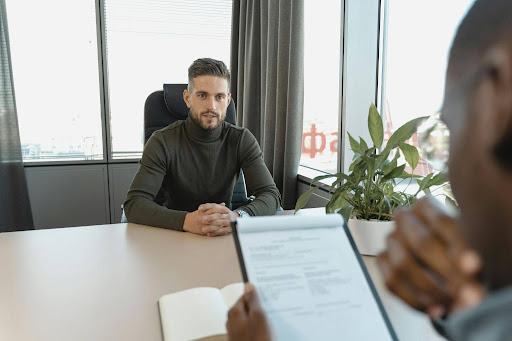A candidate being interviewed for a job by an employer, sitting opposite each other at a desk in an office