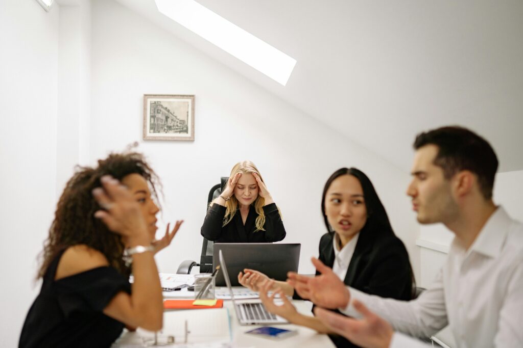 the image shows four people sitting around a table at a office workplace in distress. The two individuals in the front of the group are in an argument with the next closest person watching while the last person puts has their head lowered into their hands.