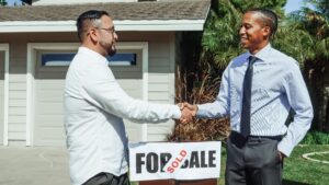 image shows two men standing outside of a house with a for sale sign that now says 'sold'