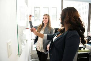 two female colleagues are working on a task using a whiteboard. They are smiling and working happily together
